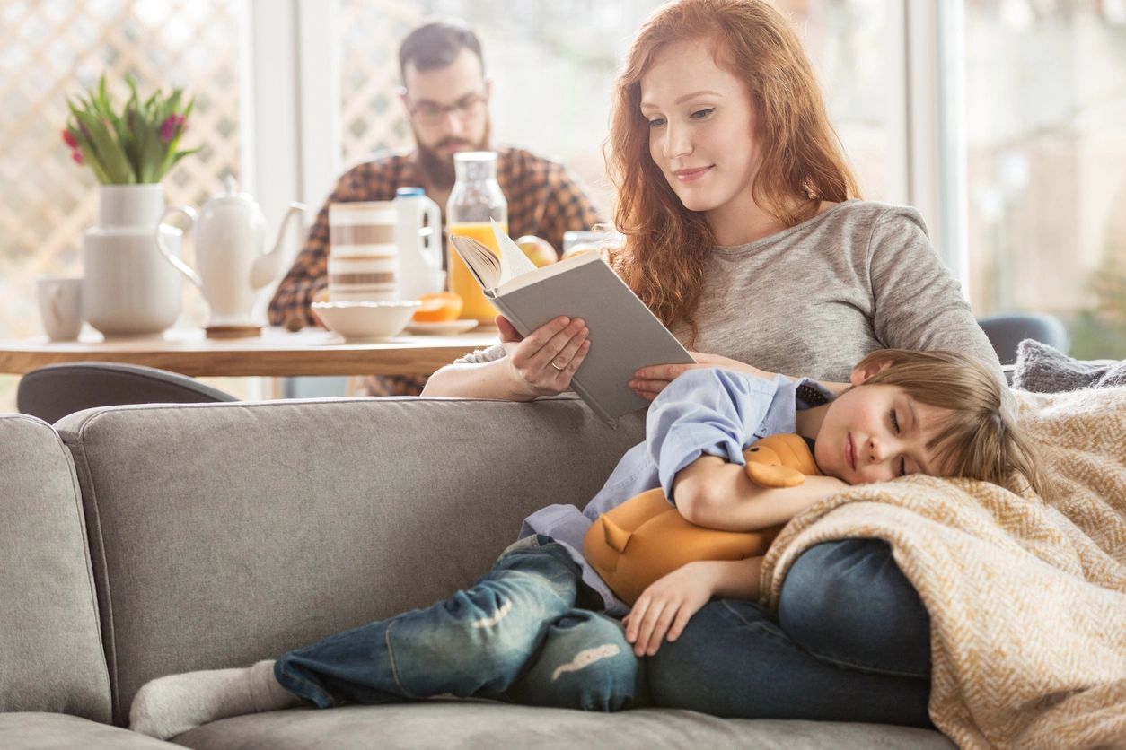 Sleeping son lying on mother's lap while she is reading a book and father eating in the background